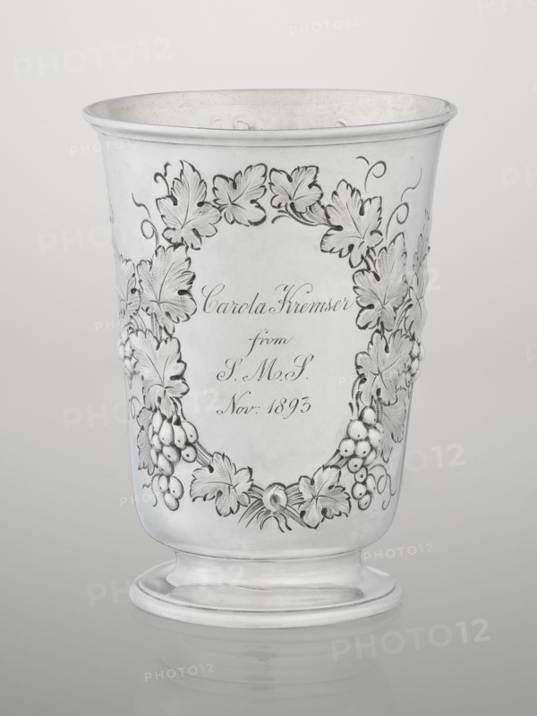 Conical silver mug on a circular foot by Barnard, Edward & Sons Ltd London; goldsmiths; fl. ca. 1680–, slightly flared profiled rim, gilded interior. Belly decorated with relief vine wreaths; one wreath bears an English inscription: Carola Kremser / from / SMS/ Nov: 1893. Date noted 1861/1862, England; height 12 cm, diameter 9 cm, weight 175 g., The image shows a metallic cup with an outward flaring rim and a rounded pedestal base, rendered in shades of silver and gray. The cup surface features an ornate raised garland of leaves and clusters of berries wrapping around the front, with textured leaf veins and curled tendrils. The interior and rim appear slightly darker silver, while the exterior is a polished light silver. The background is a smooth gradient of pale gray.