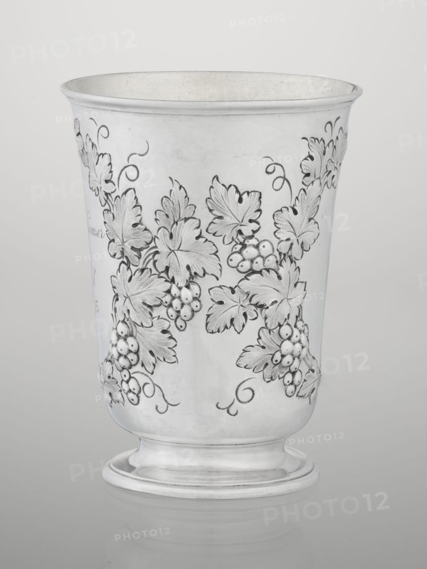 Conical mug, silver on circular foot; belly decorated with relief vine wreaths, one with an English inscription: Carola Kremser / from / SMS/ Nov: 1893. Slightly flared, profiled rim. Gilded interior. Maker: Barnard, Edward & Sons Ltd London, goldsmith's firm, active ca. 1680–. Date: 1861/1862. Place: London, England. Function: vessel. Height 12 cm; diameter 9 cm; weight 175 g. Provenance: scrap - purchase, purchase; dedications; relief decoration; vine ornament., A metal cup with a flared rim and rounded foot, finished in a reflective silver tone, decorated with an engraved pattern of vine leaves, curling tendrils, and clusters of round berries rendered in darker engraved lines and shaded areas; the surface shows subtle variations in sheen and faint wear marks, and the background is a smooth neutral gray-white.