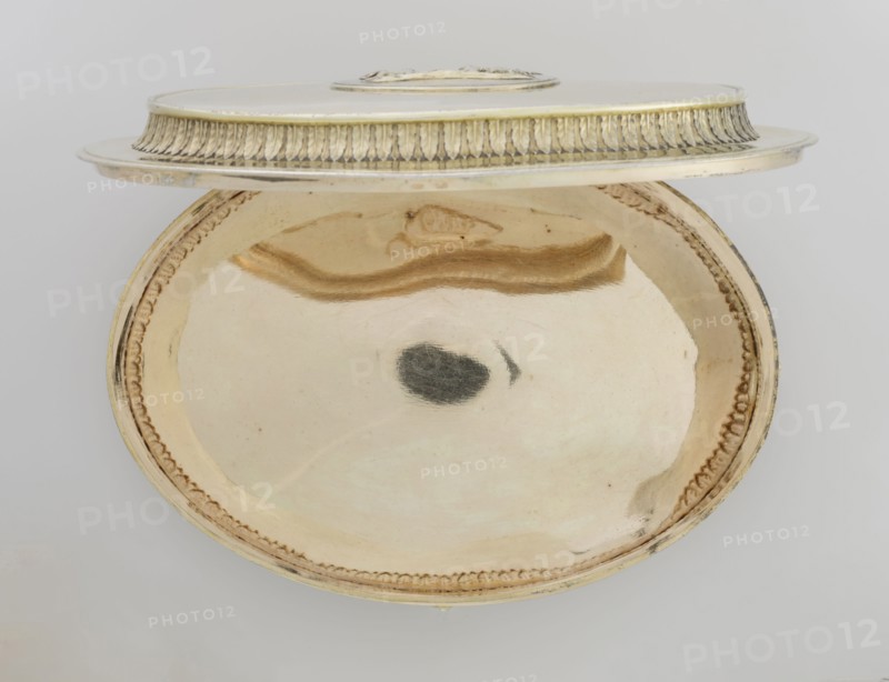 Oval toilet box by Thomas I Pitts fl. 1755–ca.1793, goldsmith. Oval box on four volute feet formed of acanthus leaves; lower edge with a band of bay leaves, above them laurel festoons with suspended medallions containing rosettes, upper edge with an anthemion frieze. Hinged recessed cover bordered with a band of bay leaves matching the body; central medallion on the cover with a relief of four putti at a sacrificial altar. Classicism/Georgian/George III style; relief decorations, festoons, palmettes, anthemions, bay leaves. Dated 1780–1781. England, London, Great Britain. Toilet accessory. Height 12 cm; width 27.1 cm; depth 20 cm; weight 1627 g., A round metallic dish with a lid shown partly open, the interior of the dish reflecting a smooth surface with a few darker smudges and a curved reflective streak; the lid has decorative ridged edging and a raised central area. The visible colors include gold, beige, cream, silver, and a few dark gray reflections against a pale gray background.