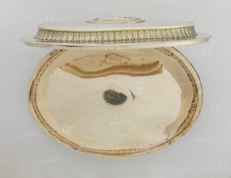 Oval toilet box by Thomas I Pitts fl. 1755–c.1793. Silver; Georgian/George III style, 1780/1781, London, England. Oval box on four volute feet formed of acanthus leaves. Lower edge decorated with a band of bay leaves; above, laurel festoons with suspended medallions containing rosettes; upper edge with an anthemion frieze. Hinged recessed cover bordered with a band of bay leaves matching the body. Central medallion on the cover with a relief of four putti at a sacrificial altar. Ornament: festoons, palmettes, anthemions, bay leaves; relief decorations. Function: toilet accessory. Dimensions: height 12 cm, width 27.1 cm, depth 20 cm; weight 1627 g., A shallow oval metal dish with a raised rim and a matching lid set slightly ajar, both showing a smooth reflective interior and decorative edging; the dish and lid are mainly pale gold and warm beige with areas of darker golden brown reflections and a small dark grayish spot near the center of the dish; the rim decoration and lid trim show a...