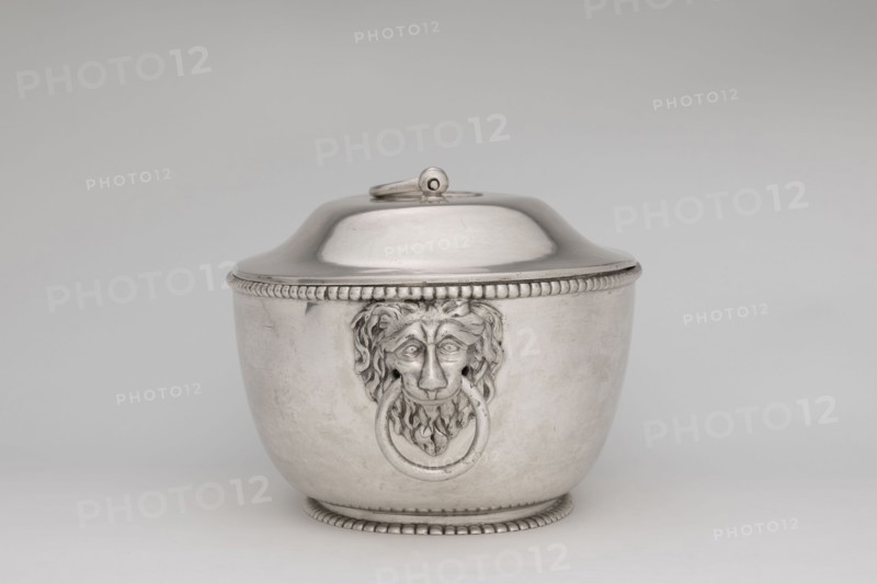 Vase with cover and platter; maker: Marcin Holke fl. ca. 1765–ca. 1792, goldsmith; tableware elements, vases; 18th century; Poland, Warsaw; vessel; height 20 cm, width 46 cm, depth 31.5 cm., A silver-colored metal lidded container with a rounded body and domed lid sits centered against a light gray background, the lid having a small looped handle on top and beaded trim around its lower edge. The body of the container features a decorative lion-head motif in the center holding a round ring in its mouth, and matching beaded trim runs along the base and top rim. The surface of the container is smooth with subtle reflections and faint scuffs, and the overall color palette includes shades of silver, gray, and off-white.