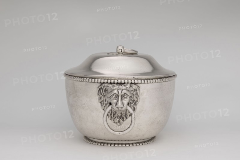 Vase with cover and platter; maker: Marcin Holke fl. ca. 1765–ca. 1792, goldsmith; tableware elements, vases; 18th century; Poland, Warsaw; vessel; height 20 cm, width 46 cm, depth 31.5 cm., A metallic container with a rounded lid and a loop handle on top sits against a plain background, the container showing a raised beaded rim near the lid and a matching beaded base; the front features a sculpted lion head with a large ring held in its mouth, and the surface shows subtle scratches and reflections. The visible colors are silver, gray, and muted off-white in the background.
