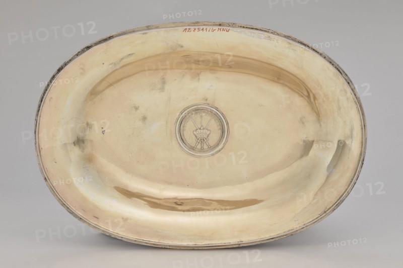 Vase with cover and platter, Marcin Holke fl. ca. 1765–ca. 1792, goldsmith; tableware elements, vases; 18th century; Poland, Warsaw; vessel; height 20 cm, width 46 cm, depth 31.5 cm, An oval shallow dish with a reflective metallic surface showing a warm yellowish-gold tone with areas of paler cream and subtle silvery highlights, a slightly darker golden-brown smudge near the lower rim, and faint grayish tarnish marks and smudges scattered across the interior; a circular raised medallion sits near the center with a central emblem in a slightly darker metallic shade, and the dish has a thin rim with worn darker gray edges.