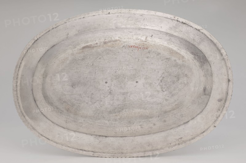 Vase with cover and platter, by Marcin Holke fl. ca. 1765–ca. 1792, goldsmith; tableware elements, vases; 18th century, Poland; Warsaw; vessel; height 20 cm, width 46 cm, depth 31.5 cm, An oval metal tray with a worn, scratched surface in shades of gray and off-white, showing areas of darker gray patina and faint brownish discoloration, a raised rim with a slightly scalloped edge, and a shallow recessed center with visible scuffs and rings.