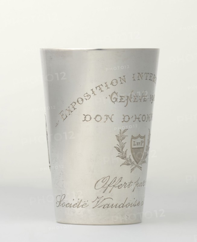Art Nouveau conical mug by Jezler & Co Schaffhausen; silversmith; est. 1822. Relief ornament of lily of the valley flowers; engraved inscription: EXPOSITION INTERNATIONALE / GENEVE 1907 / DON D'HONNEUR and cartouche with laurel branches containing monogram L. ET P. and italic Offert par la / Societe d'Aviculture. Dated 1907, Switzerland Schaffhausen. Height 9 cm, width 6 cm, weight 78 g., A cylindrical metallic cup with a smooth light gray surface stands upright against a pale off-white background, showing engraved decorative elements including a laurel-like emblem and flowing script-like ornamentation; the metal has subtle wear marks and faint scratches, and the overall tones visible are silver-gray, light gray, off-white, and a hint of darker gray in the engraved lines.