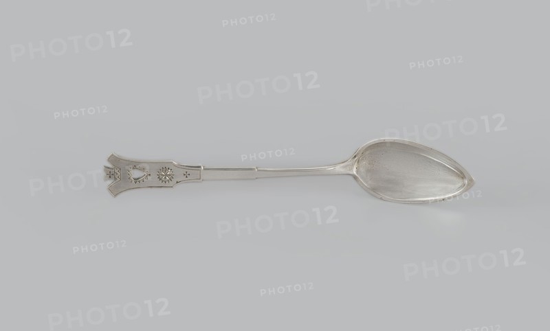 Dessert spoon, Jarra, Marcin 1852–1938 goldsmith, Zakopane style, early 20th century, Kraków, Poland. Dessert spoon with an ovoid, smooth, pointed scoop, a stepped handle ending in a stylized crown and decorated with an engraved geometric ornament stylized to resemble Zakopane style. Cutlery; height 16 cm, width 3 cm, weight 35 g., A single metal spoon lies horizontally on a light gray background, showing a reflective silver color overall with subtle darker gray tarnish spots inside the bowl and along the handle; the bowl is elongated and teardrop-shaped with a smooth interior, while the handle is flat and tapers slightly toward an ornate flared end that features engraved geometric and decorative cutout shapes; the scene contains shades of silver, light gray, and darker gray.