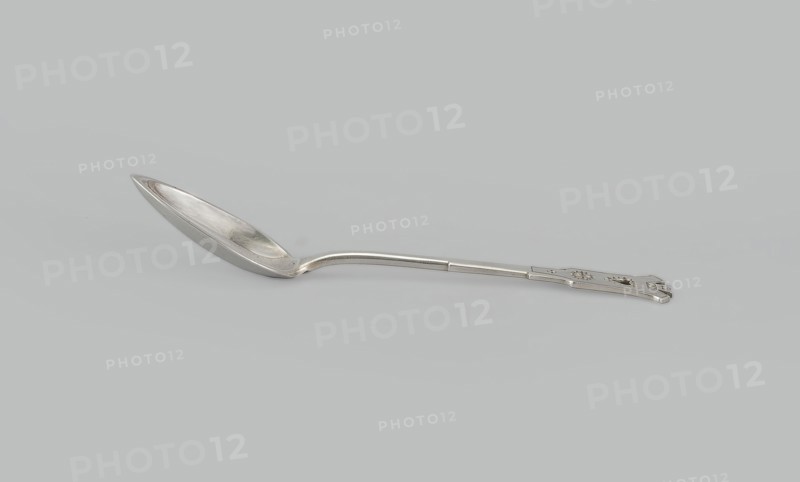 Dessert spoon, Jarra, Marcin 1852–1938 goldsmith; maker, Paderewski, Ignacy Jan 1860–1941 - testamentary bequest provenance, Zakopane style, Dessert spoon with an ovoid, smooth, pointed scoop, a stepped handle ending in a stylized crown and decorated with an engraved geometric ornament stylized to resemble Zakopane style, early 20th century, Poland, Kraków, cutlery, height 16 cm, width 3 cm, weight 35 g, A single metal spoon lies horizontally against a light gray background, showing a reflective silver surface with subtle highlights and shadows; the bowl of the spoon is elongated and slightly curved with visible wear and faint marks, the handle is straight with a gentle bend near the bowl and ends in a flat decorative section that has a few small cutout shapes and raised details, and the overall colors visible are silver and shades of gray.