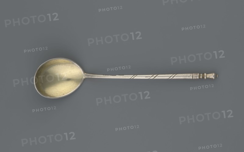 Decorative spoon, Dmitriyev, M fl. 1854–1877 goldsmith; Decorative spoon with a teardrop-shaped scoop, decorated on the reverse with a polished niello acanthus twig; angular handle at the base, becoming conical, ending with a bowl. 1863, Moscow, Russia; cutlery; height 13 cm, width 2.8 cm, weight 20 g., A single metal spoon lies horizontally against a solid muted gray background, showing a rounded bowl with a warm golden interior and a cooler silvery outer rim; the long narrow handle is silver with subtle linear grooves and a twisted decorative band near the end, which terminates in a small turned finial that is slightly darker.