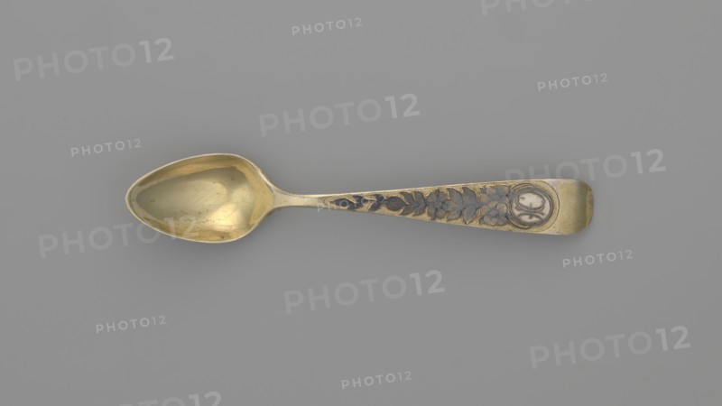 Tea spoon by Fedor Klimov Buszkowskij fl. 1795–1834, goldsmith. Decorative gilded spoon with an ovoid scoop with a pointed tip; reverse decorated with a niello ornament of fish scales and rocailles. Flat handle widening at the end, slightly bent and rounded, with niello decoration on both sides with floral motifs. Incised coats of arms. Ornament: flowers, fish scale. Date: 1815. Place of origin: Veliky Ustyug, Russia. Material/function: cutlery tea, teaspoon. Dimensions: length 14 cm, width 2.6 cm, weight 15 g., A single tablespoon with a rounded bowl and a tapered handle lies flat against a plain background; the spoon has a metallic sheen in gold and brass tones with darker grayish areas in the recessed decorative elements, and the handle features a floral and leaf pattern in contrasting darker gray set into the gold surface, ending in a rounded tip with a circular recessed area; the background is a uniform medium gray.