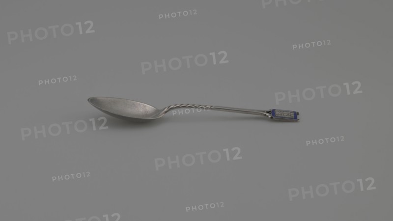 Souvenir spoon, Ford Company fl. 1875–1899. Souvenir spoon with an ovoid scoop with a pointed tip; half-handle with oblique cuts, then flat, splitting at the end, with a plaque attached in the form of a cartouche with an open book bearing the Hebrew inscription URIM/TUMMIM on a background of sapphire enamel. Inscription: Paderewski, Ignacy Jan 1860–1941. Date: 1890s. Place of origin: New Haven, United States of America. Materials/scale: cutlery; length 10.5 cm, width 2.4 cm, weight 15 g., A single metal spoon lies on a smooth surface, showing a reflective bowl with a slightly worn texture and a twisted section along the handle; the handle ends with a small rectangular decorative panel that includes a darker colored inset. The visible colors are silver, gray, and a small area of blue on the handle.