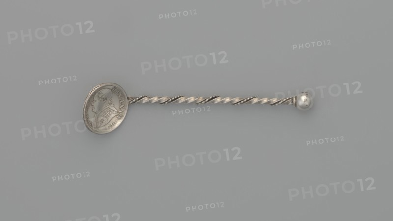 Souvenir spoon, souvenir spoon with a scoop made of a 10-soldo coin with an image of Pope Pius IX in profile on the obverse and the inscription PIVS IX PON MAX A XXII; on the reverse the inscription STATO PONTIFICO 50 R CMI and in the middle in a laurel and oak wreath - 10 / SOLDI / 1868. The handle is made of twisted wire with a ball at the end. 1868 coin, end of the 19th century, Italy. Cutlery, varia. Length 7.5 cm, width 1.8 cm, weight 5 g., A slender metallic object lies horizontally on a muted gray background, showing a twisted rod with alternating smooth and ridged spirals that have a silvery sheen; one end features a round flat disk with a raised profile of a person in relief and decorative patterns around its edge, while the opposite end terminates in a small polished spherical knob with slight surface blemishes.