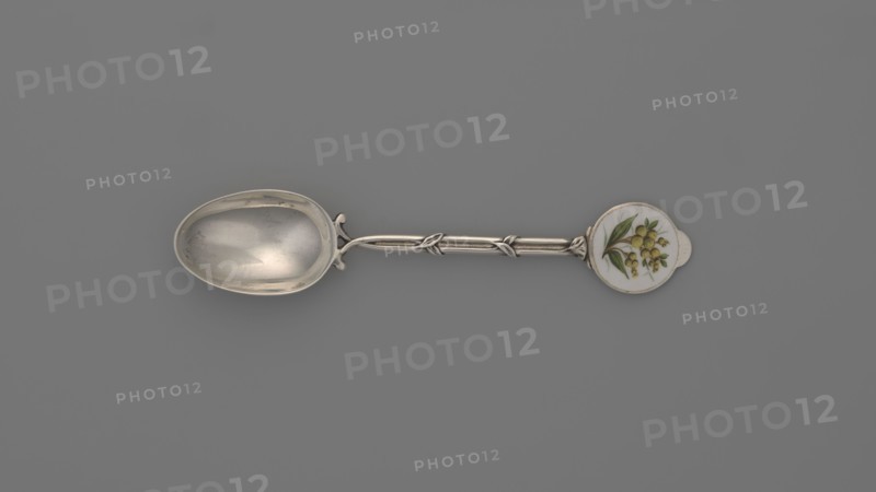 Decorative spoon by Deakin & Francis Ltd Birmingham; goldsmiths; fl. from 1786. Spoon with an ovoid smooth scoop; handle formed of two intertwined stylized branches with leaves, ending in a round flat terminal with enameled decoration depicting on a white background a branch with yellow spherical fruits and lanceolate green leaves. Inscribed/linked to Paderewski, Ignacy Jan 1860–1941. England, Birmingham, Great Britain, 1912/1913. Cutlery; height 11.5 cm, width 2.4 cm, weight 15 g., A silver spoon laid horizontally on a gray background with an oval bowl on the left showing reflective silver surfaces and subtle scratches, a decorative silver handle with twisted and knotted elements in the middle, and a round decorative end on the right featuring a white circular inset painted with a botanical motif of green leaves and small yellow berries; visible colors include gray, silver, white, green, and yellow.