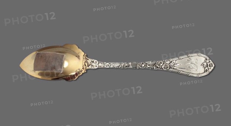 Dessert spoon, Durgin Co Concord; goldsmith's firm; fl. 1853–1924. Dessert spoon with an ovoid scoop topped with a pointed tip, decorated with semi-plastic roses at the bottom, gilded; handle widening towards the end and topped in a semicircle, edged with a profiled band, decorated with roses. Dauphin pattern introduced by the company in 1897; 1899–1905 the HP — Helena Paderewska — monogram indicates the spoon was purchased or given no earlier than 1899, the Paderewskis' wedding date. Paderewski, Ignacy Jan 1860–1941 and Paderewska, Helena 1856–1934 monogram. United States of America; Concord. Cutlery. Length 15 cm; width 2.8 cm; weight 30 g., A single ornate spoon lies against a flat gray background, showing a polished golden-toned bowl with a small rectangular patch of coarse fabric attached inside, and a decorative silver-toned handle featuring floral and scroll motifs running along its length and into an embellished terminal with floral accents; visible colors include gray, gold, s...