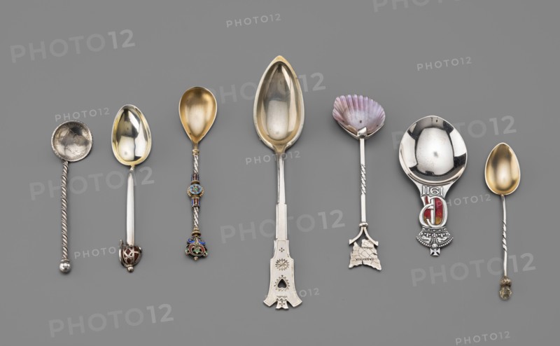 Souvenir spoon by Fenton, James Birmingham; goldsmith's firm; fl. 1852–1954. Spoon with an ovoid scoop gilded inside, angular handle ending with an openwork sword hilt set with an agate. Late 19th century, Birmingham, England. Cutlery; height 10.5 cm, width 2.8 cm, weight 10 g. Paderewski, Ignacy Jan 1860–1941 mentioned in association; decorative stones, swords., A row of several decorative spoons is arranged horizontally on a gray background; the spoons show a variety of shapes and embellished handles. Visible colors include silver, gold, purple, red, blue, green, and brown. Some spoons have plain oval bowls with a gold-toned interior, one spoon has a scallop-shaped bowl with purple and pink tones, and another has a deep reflective silver bowl. The handles vary: a few are twisted silver, one has colorful enamel segments in blue and green with gold accents, one handle ends in a flat decorative plate with cut-out shapes, and another features a rounded decorative finial with red enamel. ...