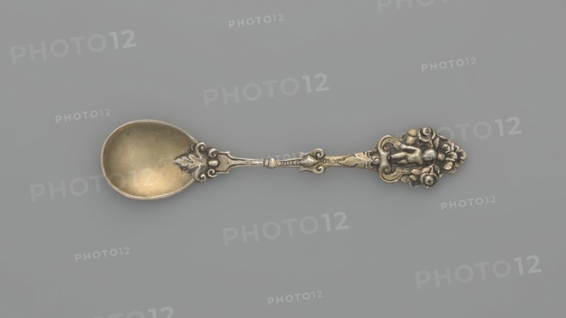 Decorative spoon, Neresheimer Hanau; goldsmith's workshop; 1890-, goldsmith's company author, Paderewski, Ignacy Jan 1860-1941 - testament provenance, grotesques, neo-Renaissance style, putti. Decorative spoon with a circular scoop and a profiled handle topped with a grotesque with a putti standing in an arcade decorated with roses, late 19th century, Germany, Hanau, cutlery, varia; length 11 cm, width 2.4 cm, weight 20 g, A single ornate metal spoon lies horizontally on a flat background, showing a bowl with a slightly worn golden tone and a decorative handle with intricate raised floral and scroll motifs in a darker silvery and warm brass color; the background is a smooth muted gray.