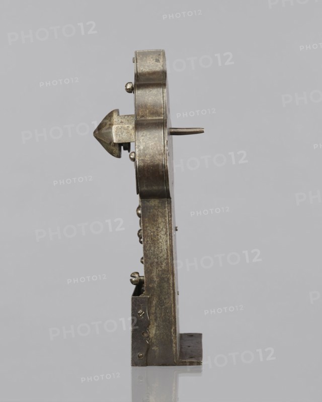Four-bolt door lock with key. A lock of good locksmithing workmanship, from the beginning of the last century, with precise, professional finishing on both the lock plate and the brass covering of the mechanism. The key originally and its rich register are meticulously and precisely crafted, showing a high level of locksmithing skill—probably a masterful piece of craftsmanship. A four-bolt latch lock on a rectangular plate with a trefoil rear section, with a cover engraved with a twig motif, profiled male busts with mustaches in the side bulges and an en face mask in the central one, and a hole for the register dome. On the reverse, the plate is bent at a right angle for mounting on the side of the door; the mechanism is partially covered by an openwork cover engraved with a twig motif with a bird and a pomegranate. Screw mounting. The key is missing, and a brass box cover that once covered the central part of the lock is also absent. 17th century, Baroque style, metalwork; height 33 c...