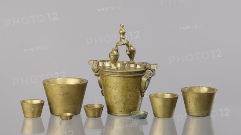 Set of weights, Seven weights in the form of bowls—truncated cones—and an eighth, largest piece serving as a container for the others, with a hinged lid closed by a hasp and a movable handle mounted in pinnacles shaped like simplified vases. On the lid: 16 MARK and a Prussian eagle; hallmark BERLIN; another mark engraved 160; 152 engraved on the side weight 1865.57 g. 18th century, Germany. Weight; height 17.5 cm, width 10.2 cm, depth 14.3 cm., A group of metal vessels arranged on a reflective surface with a plain light gray background, including a central ornate vessel with a decorative handle and two small finials, flanked by several simpler cup-shaped vessels of varying sizes and shapes. The objects show a metallic gold color with areas of darker patina and small spots of greenish corrosion on one smaller piece, and faint reflections of the items appear on the surface beneath them.