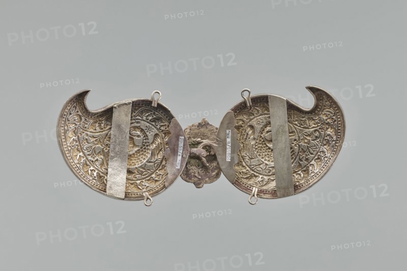 Buckle, Repossession; gift. Silver buckle fastening Delya's coat, probably royal, inscription 24 February 1705. Two-part, each element in the form of a bota, with a repoussé border composed of strips of pearls and leaves. Inside, circular medallions with a phoenix holding snakes in its beak. Both elements are connected with a hook, the part with the ring covered with a rectangular shield with ogee spines at top and bottom, bearing a circular medallion with the Polish eagle. Both parts at top and bottom have soldered rings; on the reverse, vertical strips for attaching a belt are soldered on; one of them with a disc on the clasp is engraved in italics with the words Warszawa / 24 11 1705. Not signed, not hallmarked. The buckle may be a Middle Eastern work from the 19th century, later supplemented with a disc with the Polish eagle and an inscription, probably from the third quarter and made in Europe. 3rd quarter of the 19th century; clothing accessory buckle; height 10.5 cm, width 26.5 ...