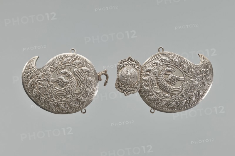 Buckle, repossession, gift. Silver buckle fastening Delya's coat, probably royal, inscription 24 February 1705. Two-part buckle, each element in the form of a bota, with a repoussé border composed of strips of pearls and leaves. Inside, circular medallions with a phoenix holding snakes in its beak. Both elements are connected with a hook; the part with the ring is covered with a rectangular shield, with ogee spines at the top and bottom, bearing a circular medallion with the Polish eagle. Both parts at the top and bottom have soldered rings; on the reverse, vertical strips for attaching a belt are soldered on; one of them with a disc on the clasp is engraved in italics with the words Warszawa / 24 11 1705. Not signed, not hallmarked. The buckle may be a Middle Eastern work from the 19th century, later supplemented with a disc with the Polish eagle and an inscription, probably from the third quarter and made in Europe. 3rd quarter of the 19th century; Germany. Clothing accessory buckle....