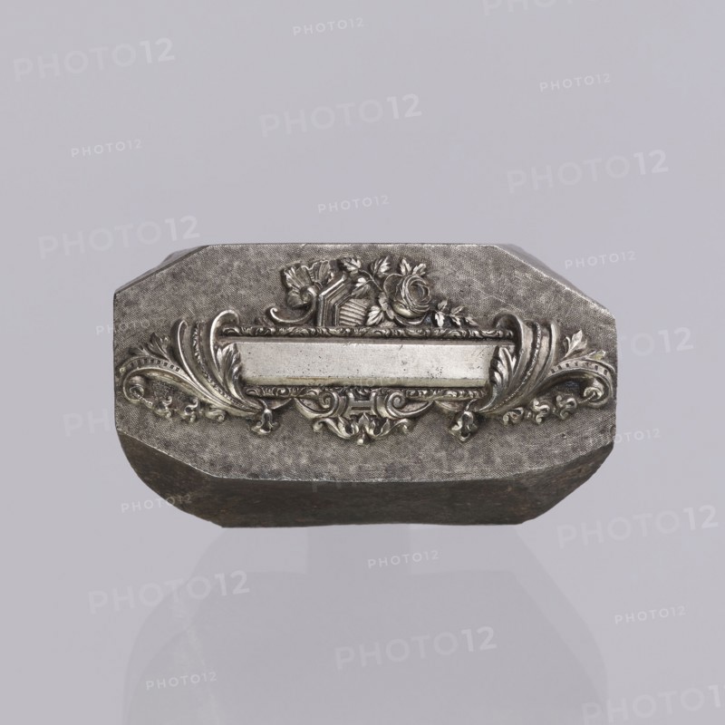 Jewellery die, Approximately hexagonal die with a relief rectangular reserve enclosed at the ends by two leaves. At the top and bottom of the reserve are floral and plant decorations and scrolls. Probably a jeweller's die for making semi-finished products, 2nd half of the 19th century, Germany. Die; height 3 cm, width 6 cm, depth 3 cm., A metallic, octagonal object sits against a pale gray background, showing a raised, ornate central plaque with a horizontal blank panel flanked by symmetrical scrollwork and leafy flourishes; above the panel is a decorative motif including a rose and ribbon-like elements, and the metal surface displays a textured, slightly worn finish with darker areas along the lower edge.