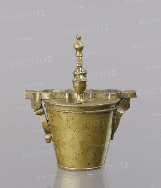 Seven weights in the form of bowls—truncated cones; the eighth, largest, serves as a container for the others and has a hinged lid with a movable handle mounted in pinnacles shaped like simplified vases. On the lid: stamped 16 MARK, Prussian eagle, BERLIN stamp; another mark engraved 160; on the side engraved 152. Late 18th century, Germany. Materials: measures and weights. Maker: unknown., A brass container with a domed lid and a turned finial on top, showing a warm golden color with areas of darker patina and small spots. The container has a tapered cylindrical body and a decorative rim beneath the lid with a subtle rope-like pattern, and a pair of hinged bracket-like handles on either side with rounded end pieces, all in the same brass tones. The lid has an irregularly shaped flat top section with cutouts around the central finial, and the finial itself is a turned, vase-like form stacked with rounded and cylindrical segments. The background is a uniform light gray and the object re...
