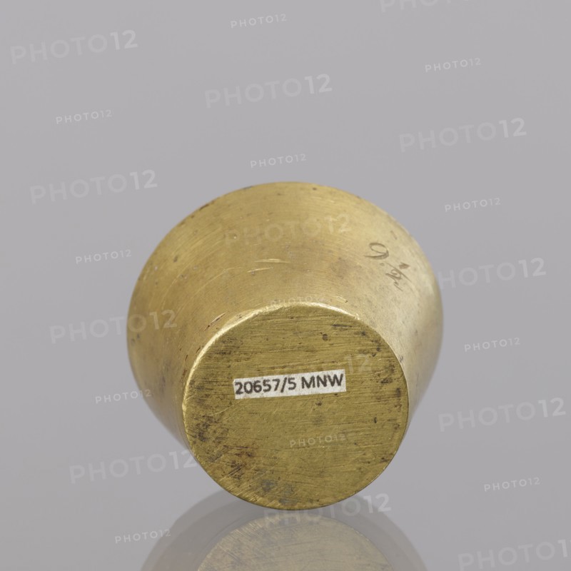 Gwicht;, In the shape of a truncated cone; engraved marking on the side 91/2, inside 8, eagle; 116.45 g; end of 18th century; Europe, Germany; weight; height 2.9 cm; width 4.5 cm., A cylindrical metal object viewed from one circular end, showing a worn, slightly rough surface with scratches and small dark spots; the metal has a yellowish-gold color with areas of darker discoloration and some lighter highlights; it rests on a smooth, reflective gray surface that creates a faint reflection below; the background is a uniform pale gray.