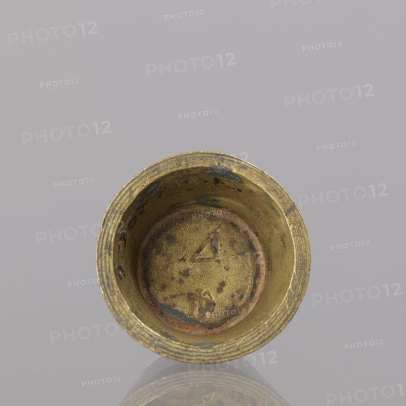 Gwicht, unknown creator; Szewczykowski, Roman 1849–1901. Measures and weights: in the shape of a truncated cone; markings on the side engraved 43/4, inside 4, eagle; 57.84 g; 18th century; Europe; Germany; height 2 cm; width 3.8 cm., A close-up top view of a cylindrical metal cup with a weathered golden surface showing patches of greenish-blue and reddish-brown discoloration, a textured rim with concentric grooves, and a slightly mottled interior base; the background is a smooth light gray.