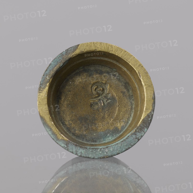 Gwicht, unknown, Szewczykowski, Roman 1849-1901 — In the shape of a truncated cone, internal marking 2, weight 24.75 g, 18th century, Europe, Germany, height 1 cm, width 3.1 cm, A round metal object with a shallow recessed interior, showing worn surfaces and an indented decorative mark in the center; the rim is thick and uneven with areas of wear and patina. The visible colors include brassy gold, dark brown, greenish-blue verdigris, and muted gray tones. The object sits against a light gray background and its underside is reflected faintly on a glossy surface below.