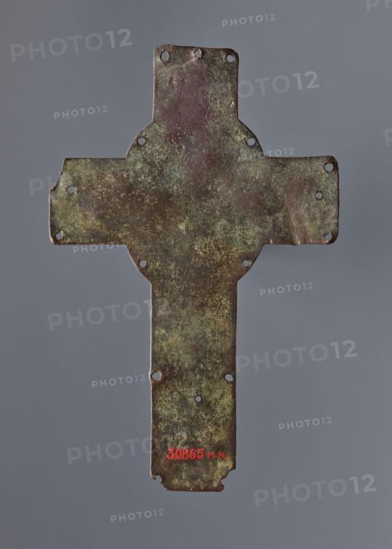 Cross from the binding of a liturgical book or processional, liturgies, processions, enamelling, Romanesque cross with a relief image of Christ and the inscription XPS/IHS. Originally enameled, currently without traces of enamel. Only the feet of the Crucified figure remain. Found in 1911 in a field near Slomniki then within the Kingdom of Poland. Described as a plaque from the binding of a liturgical book; possibly a processional cross. May come from Kacice, which belonged to the Cistercians who later settled in Mogila. Beginning of the 13th century, Limoges, France. Cross, height 21 cm, width 13.5 cm., A weathered metal cross-shaped plate with irregular edges and a group of small circular holes near the ends and along the body, showing a mottled surface with patches of green, brown, dark reddish-brown, and hints of yellowish discoloration, set against a plain neutral gray background.