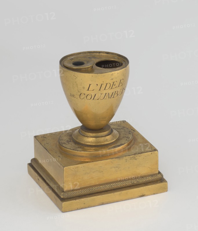 Inkwell, early 19th century, France, maker, attributed to Tadeusz Bylewski 1866–1939; inkwell; height 13.5 cm, width 9.4 cm, depth 8.4 cm., A small metallic object sits on a flat surface, composed of a rounded cup-like top mounted on a stepped square base. The cup portion has two round openings at its rim and shows a smooth, reflective surface with some scuffs and dark spots. The base is tiered with a textured band around its lower tier and displays worn areas and surface marks. The overall color palette is primarily gold with darker tarnished areas and small blackish spots, set against a pale gray background.