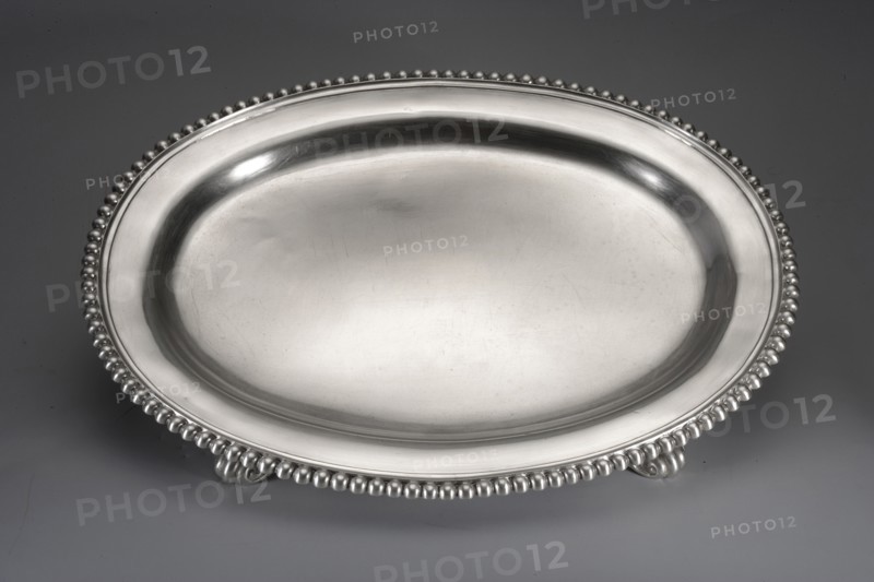 Oval tray by Jacob Haase 1704–1762, goldsmith active from 1730; died 1762; widow active to 1774. Smooth oval tray with a raised collar bordered with pearling, supported by four acanthus legs feet not original, soldered on in the 19th century. Scrap — purchase; purchase provenance. Ornament: pearling and acanthus leaves. Dated 1750, Gdansk, Royal Prussia, Poland. Vessel. Height 3.7 cm; width 35 cm; depth 27.2 cm; weight 1035 g., A large oval metal tray with a smooth reflective surface and a raised rim decorated with a beaded edge rests on a flat surface, showing a few small ornamental feet beneath the rim; visible colors include silver tones on the tray and a muted gray background.