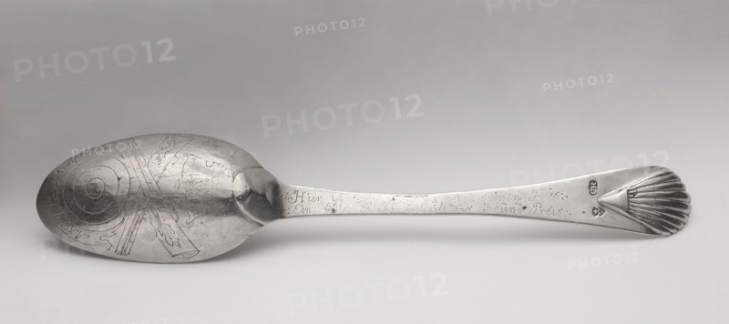 Table spoon with shooting brotherhood emblem, maker: Hermann, Christoph fl. ca. 1740–1778 goldsmith, active from the 1740s, died 1778. Silver spoon, ca. mid-18th century. On the reverse of the bowl: dotted italic inscription Anno 1766 1 April; below it an engraved shooting shield, crossed rifles, and the Elblag coat of arms. Along the handle: dotted, very faded German italic inscription Hier isst ? der Platz da zeigt deinen Fleis / Ein Edlen Rath # dir Ehr und Preis. Obverse of the handle has a later inscription: AW CH / 1860 / 14 Sept. Function: shooting-award spoon awarded at annual marksman fraternity competitions. Place: Elblag, Royal Prussia. Cutlery; length 22.5 cm; weight 64 g., A single metal spoon lies horizontally on a light gray surface, showing the bowl on the left and the handle extending to the right; the bowl has engraved concentric circular patterns and decorative engraved motifs with visible scratches and a slightly worn, reflective silver-gray finish, while the handle...