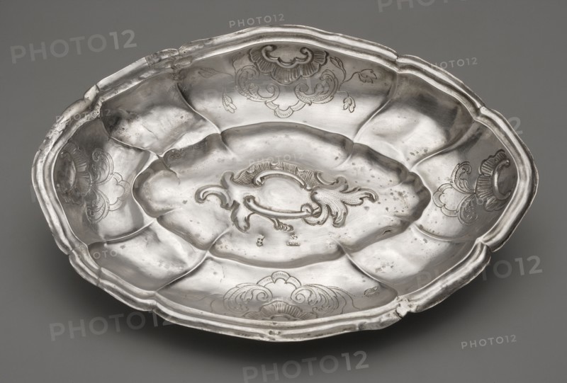 Oval cake stand, maker: Johann Constantin Lange 1702–1761; silver; rocaille and acanthus ornamentation, rooster's comb motifs. Oval silver cake stand with multi-leaf cut, set on four legs in the form of palmettes; a concave-convex mirror with a multi-leaf outline and rocaille decoration, a wavy collar vertically divided by ribs rising upwards, decorated on the longer and shorter axes with four rooster combs and an acanthus twig, and a stepped-profile rim with a multi-leaf outline. Trays of this type, popular in Gdansk goldsmithing of the period, may have been used for serving spoons. Ca. 1763; Gdansk, Royal Prussia Poland. Vessel; weight 130 g; height 4 cm; width 14.8 cm; depth 21.1 cm., A shallow oval metal dish with a scalloped rim sits against a neutral gray background; the dish has a reflective silver surface with areas of darker tarnish and small spots, a central raised decorative cartouche with flowing scroll-like motifs, and a few engraved floral or shell-like designs around the...