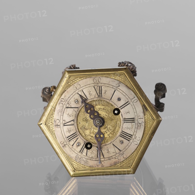 Tiled clock by Michael Gugenmus Johann Michael Gugenmus 1705–1775, watchmaker. Rococo, Regency ornament. Hexagonal table clock of gilded bronze on six volute legs with glazed side walls; silver-plated ring-shaped dial with black Roman numerals for hours and Arabic minutes, two winding holes; center engraved Regency-Rococo cartouche signed Gugenmos/Varssavie also given as Johann / Michael / Gugenmus / A Varsovie. Hinged flap closing the mechanism; front plate with a modest bun and a hammer with engraved bronze head. Mechanism with strike repeater and a handle on the side wall under the upper edge. 2nd half of the 18th century. Poland, Warsaw. Timepiece. Height 8.5 cm, width 13.5 cm, depth 13.5 cm., A hexagonal metal clock face with a brass-colored frame sits centered against a light gray background, showing a circular dial with a worn, pale surface and ornate dark metal hands. The central medallion is a warmer gold tone with engraved decorative patterns, surrounded by an outer ring that...