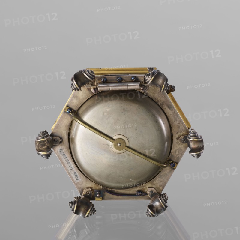 Tiled clock, Gugenmus, Michael 1705-1775 watchmaker, rococo style, regency ornament, tiled clock, table clock, hexagonal, gilded bronze on six volute legs, glazed side walls; silver-plated ring-shaped dial with black Roman numerals for hours and Arabic minutes, with two winding holes; center engraved Regency-Rococo cartouche with signature Gugenmos/Varssavie; hinged flap closing the mechanism; on the front plate of the mechanism a modest bun, a hammer with an engraved bronze head. Signature: Johann / Michael / Gugenmus / A Varsovie. Mechanism with a strike repeater, with a handle on the side wall under the upper edge. 2nd half of the 18th century, Poland, Warsaw. Timepiece; height 8.5 cm, width 13.5 cm, depth 13.5 cm., A metallic hexagonal object with a circular domed center held by a diagonal bar, showing a mix of brass, silver, bronze, and darker tarnished metal tones; each corner of the hexagon has a rounded ornamental knob with layered ridges, and several small screws and hinge-lik...