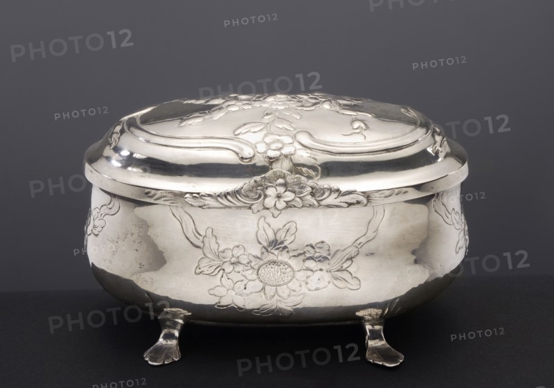Sugar bowl by Hermann II Neupert fl. 1762–1775, rocaille ornament, rococo style; egg-shaped sugar bowl on four legs shaped as stylized lion's paws; belly and cover decorated with semi-plastic bouquets of flowers and rocailles; dated 1763–1775, Berlin, Germany; silver vessel, height 8.6 cm, width 13.5 cm, depth 8.7 cm, weight 255 g., A polished metal box with a domed lid sits on a dark surface against a dark gray background; the box has an ornate floral relief design on the lid and body, curved decorative edges, and stands on a group of small sculpted feet. The visible colors are silver, dark gray, and black.
