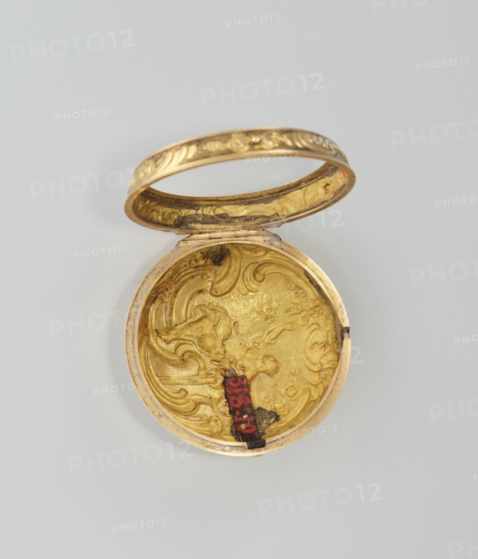 Pocket watch in case; maker: M. Cradelle fl. ca. 1750; watchmaker. Mid-18th century. London, England. Rococo style, mythological scenes. Watch in double case and outer case covered with lizard, with riveted rosette. Timepiece. Height 6 cm; width 4.5 cm; dial diameter 3.5 cm., A small round hinged container shown open, with an ornate interior and lid in metallic gold tones; the interior surface has raised swirling decorative patterns and areas of darker tarnish, and there is a small vertical strip of red and black attached near the inner edge; the exterior rim and lid edge show a gold finish with engraved decorative motifs; the background is a plain light gray.