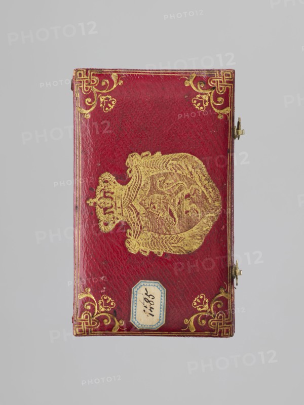 Chamberlain's key case, Rectangular key case with a hinged cover and two clasp hooks. Trimmed with red leather, with gilded embossing; on the cover a frame along the edge with an ornament in the corners. In the centre a ducal mitre. On the underside a gilded frame along the edge, in the centre an ungilded embossed rhombus with an inscribed quatrefoil, and in the centre a rhombus with a four-petal rosette. Glued on a printed card with inventory numbers numbers added in ink. 1833, Germany. Varia. Height 9.8 cm, width 5.9 cm, depth 1.8 cm., A rectangular object with a textured red surface lies against a pale gray background, decorated with ornate gold embellishments including corner flourishes and a central gold emblem that resembles a crowned shield with intricate linework; a small metal clasp is visible on one side and a small decorative label with a blue border is attached near one edge.
