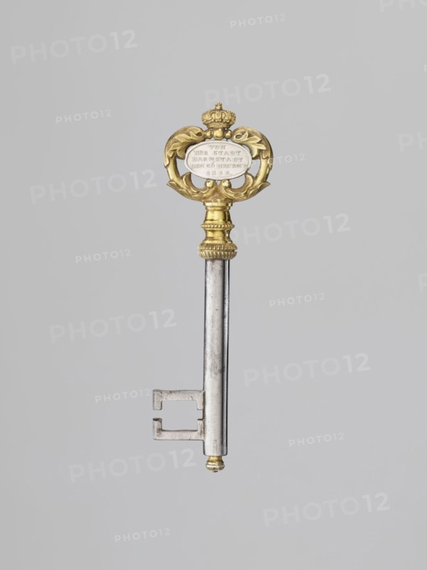 Souvenir key, steel, with a full shank and a simple feather in the form of two square brackets. Gilded bronze knot and handle: oval handle composed of two U-shaped sections decorated with leaves, fastened with a ducal mitre. Inside, an oval plaque with a Gothic monogram M and blue enamel on one side, and a cast inscription on the other: VON / DER STADT / DARMSTADT / DEN 26 DECEMB: / 1833. Given to Duchess Mathilde Caroline of Bavaria 1813–1862 and her husband Ludwig III on the occasion of their wedding, 1833. Germany; Darmstadt. Varia. Height 8.9 cm, width 2.6 cm., A single ornate key is pictured against a plain light gray background; the key has a long metallic silver shaft with a rectangular bit at the lower end and a small rounded tip with a touch of gold, and the upper section features a decorative gold head with curved foliate shapes and a small crown-like element, with accents of white visible within the central area of the head.