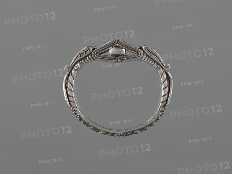 Bracelet;, scrap-purchase provenance; Chinese/Mongolian; 19th century, China, Mongolia ; materials: metal jewelry; design: dragons, dragon with pearl, hundred antiquities baigu ornament; function: body/hand ornament, fashion accessory; dimensions: height 8.0 cm, width 9.0 cm, weight 281 g., A metallic circular bracelet with ornate carved details forming a symmetrical design, showing leaf-like motifs and curved ridges around its band; a raised decorative element sits at the top center within a triangular frame. The object appears in shades of silver and gray against a uniform gray background.