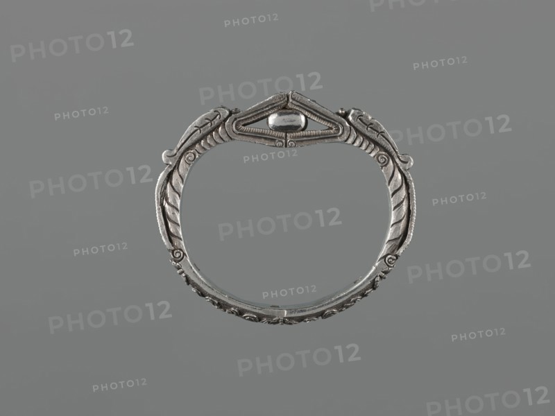 Bracelet, scrap-purchase provenance; Chinese/Mongolian body ornament hand ornament, jewelry, 19th century ; dragon with pearl motif; hundred antiquities baigu ornament; materials not specified; height 8.0 cm, width 9.0 cm, weight 281 g., A circular metallic bracelet with ornate carved detailing sits centered against a plain gray background; the metal appears silver with darker gray patina in recessed areas, featuring symmetrical leaf-like motifs and curved ridges along the band, a raised central decorative element at the top with a smooth oval bead, and fine linear textures and scrollwork around the edges, all shown in shades of silver and gray.