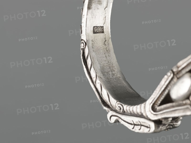 Bracelet, scrap - purchase, purchase provenance; Chinese and Mongolian art; body/hand ornament jewelry, fashion accessory; dragon with pearl motif, hundred antiquities baigu ornament; 19th century, China, Mongolia ; height 8.0 cm, width 9.0 cm, weight 281 g, The image shows a close-up of a curved metallic object with ornamental raised patterns and scroll-like details, presented against a smooth grey background; the metal appears silver with areas of darker tarnish and subtle reflections, and there is a rounded bead-like element visible at one edge.