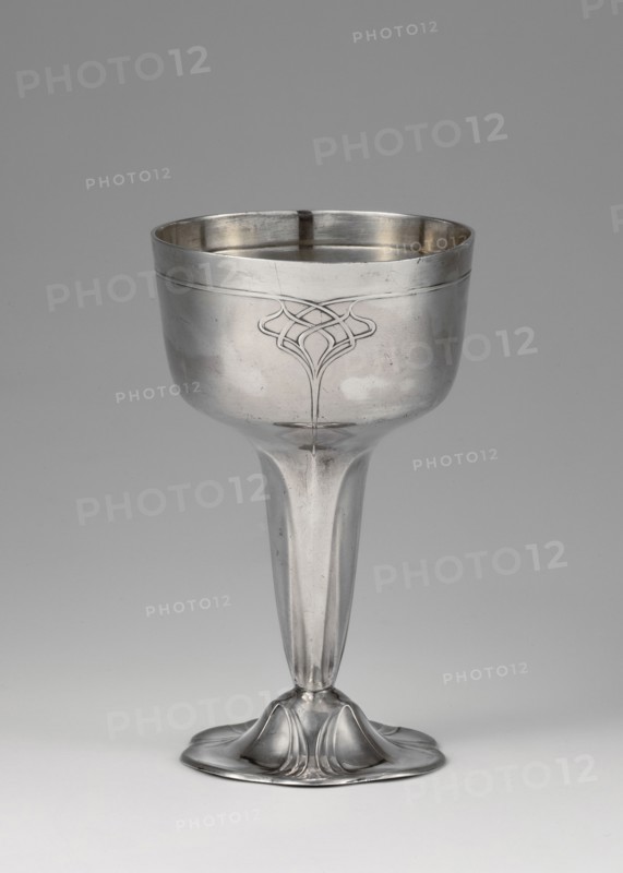 Art Nouveau silver cup by Orivit AG Cologne; goldsmith's manufactory; 1894–1905; 1906–1914 manufacturer; after takeover by WMF. Art Nouveau style; scrap. Silver Art Nouveau cup supported on a flower-shaped foot. Tall stem widening toward the top, hemispherical bowl with a slightly flattened bottom, decorated with a geometric-floral ornament. Dated 1906–1914, Cologne, Germany. Vessel. Height 12 cm; width 7.2 cm; weight 123 g., A metallic goblet photographed against a plain background, showing a bowl with a subtle engraved decorative motif on its front, a smooth tapering stem, and a rounded flared base with petal-like sculpted forms; visible colors include silver tones on the goblet and shades of gray in the background and surface reflections.