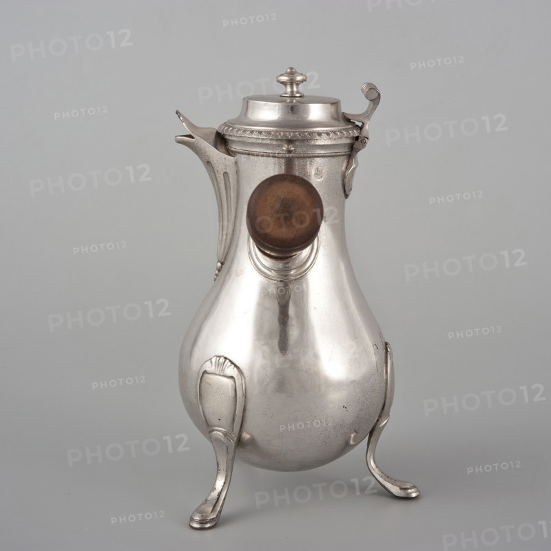 Coffee pot by F. Picard fl. ca. 1798–ca. 1809. Classification: vessel; function: coffee pot. Materials/objects: coffee pot, tableware item, jug. Date: 1798–1809. Place of origin: Paris, France. Dimensions: height 46.0 cm; width 8.5 cm; depth 8.5 cm; weight 248.2 g., A polished metal pitcher with a rounded pear-shaped body stands on a group of curved legs; the metal is primarily silver with a slightly aged patina and subtle darker spots. A short spout extends from one side near the top, and a hinged lid with a decorative finial sits atop the pitcher. A wooden knob in a warm brown tone is mounted on the front. The surface shows smooth reflective highlights and soft shadows, and the background is a uniform light gray.