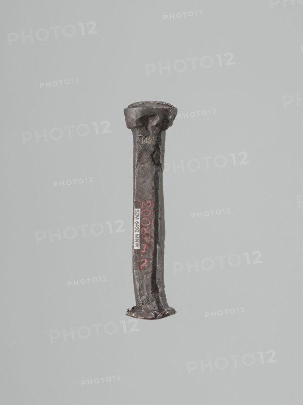 Stamp - die, Szewczykowski, Roman 1849–1901 — gift; locksmithing. Oval convex stamp-die with a depiction of the king's bust in an open crown. Origin uncertain; possibly Poland; 18th or 19th century. Die: height 9.5 cm, width 2.3 cm, depth 1.8 cm., A single vertical metal object with a rough, irregular surface and a flattened head and foot, shown against a plain light gray background, with colors including dark gray, rust-brown, black, and hints of red.