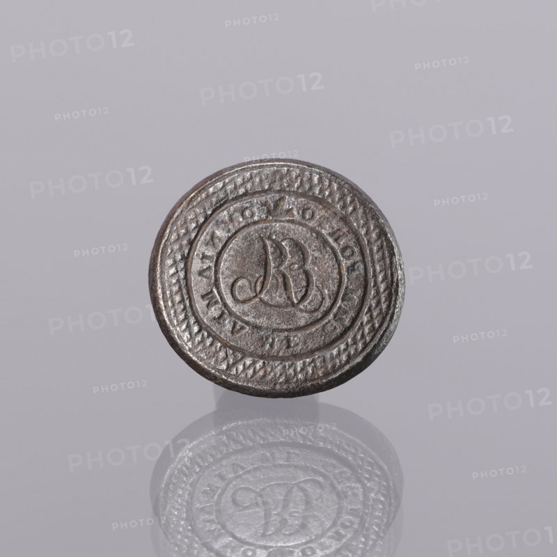 Seal stamp, engraving. Oval, almost circular; central field with engraved monogram ER R mirror-reversed. Rim surrounding central field embossed with punches reading IMMPORTO image of a bird in flight OMNIA MEA MF. Outer rim engraved with a diagonal grid. Stamp in the form of a plate with a cylindrical sleeve on the reverse for mounting a wooden handle. Early 19th century, Europe. Height 3 cm, width 3.2 cm, depth 2.4 cm., A round metallic object with a textured rim and a circular raised design in the center sits against a pale gray background; the metal appears dark silver and brownish with a slightly worn surface, and a soft reflection of the object is visible below it showing similar dark silver and gray tones.