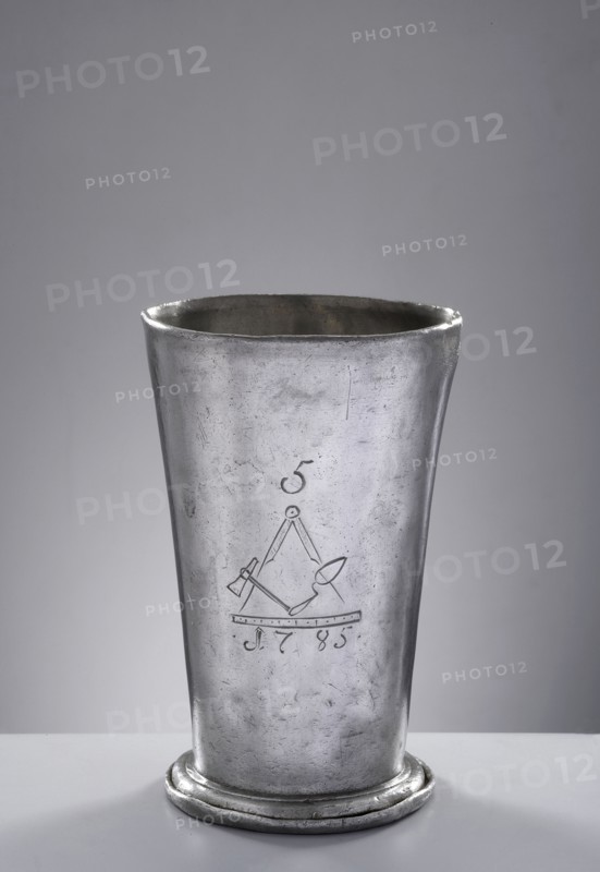 Bricklayers' guild mug. Tin mug of conical shape, set on a profiled convex base with a turned-down rim, with a smooth casing; in the center masonry emblems: compass, trowel, hammer, and square with the number 5 above and the date 1785 below. Circa 1785. Poland, Royal Prussia, Chojnice . Guild memorabilia; vessel. Diameter 9.8 cm; height 15.5 cm., A tall metallic cup with a slightly flared rim and a rounded base sits on a flat surface; the cup is silver-gray in color with a worn, scratched texture and subtle darker gray areas, and the background is a smooth, light gray gradient.