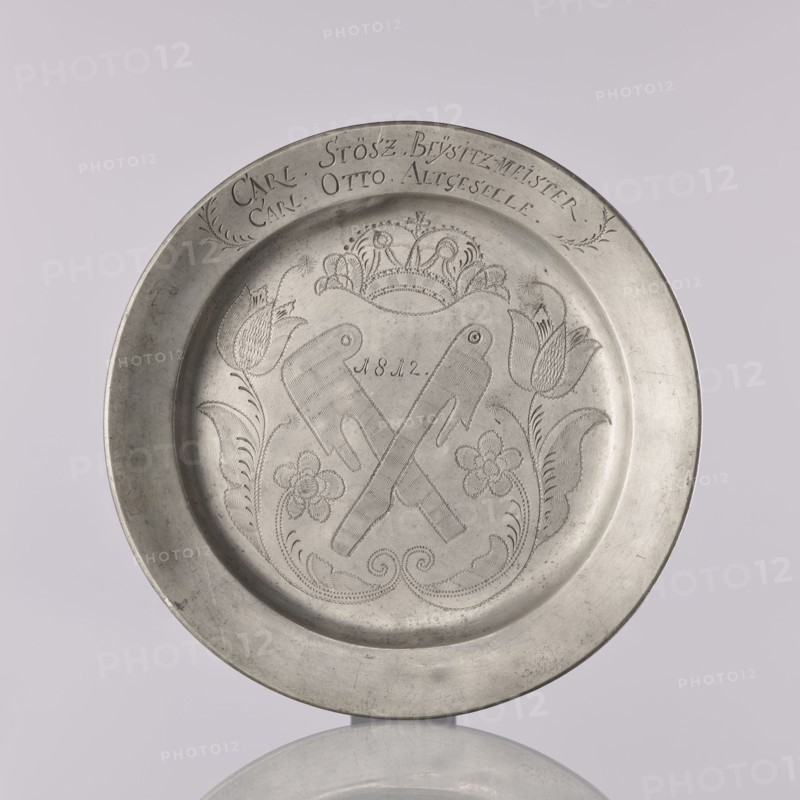 Plate by Otto Friedrich Groth fl. 1792–1822. Flat pewter plate with a smooth mirror, semicircular notch, and narrow upturned collar. In the mirror is an engraved representation of unidentified crossed guild tools with the date 1812 possibly meat cleavers, surrounded by a stylized plant twig with symmetrically arranged flowers, including tulips on leafy stems, beneath a crown. On the collar is an engraved inscription surrounded by leafy branches: CARL. STOSZ. BEYSITZ-MEISTER. / CARL OTTO ALTGESTELLE., 1812. Brzeg, Silesia. Plate; height 2.2 cm, width 22.2 cm., A round metal plate with a raised rim, shown against a pale background, featuring an engraved central design of two crossed objects surrounded by floral motifs and decorative scrollwork, with a crown-like element above and stylized flowers and leaves flanking the central motif; the plate has a satin metallic surface with areas of darker and lighter gray.