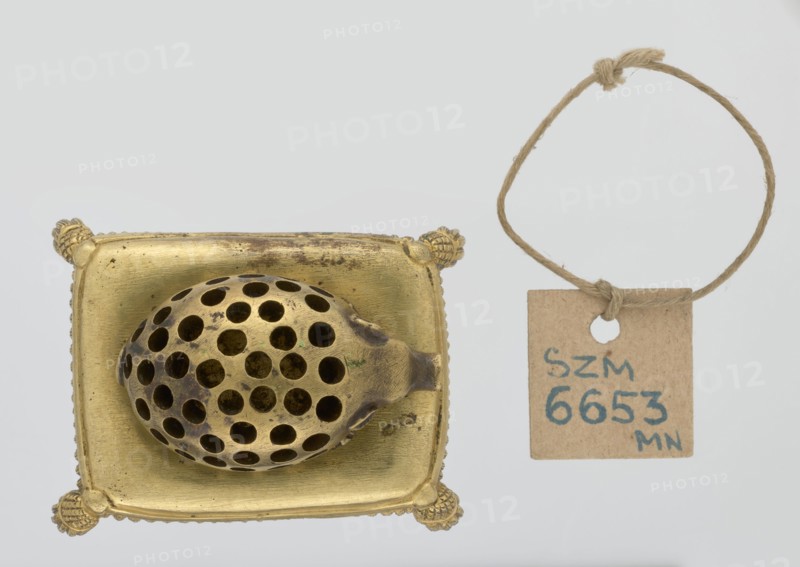 Toothpick holder, hedgehogs, animals, tassels. Bronze toothpick holder in the shape of a pig resting on a rectangular base bordered with a fringe. 1820s–1840s, Poland . Varia. Height 3.5 cm; width 6.3 cm; depth 4.7 cm., A metallic object with a golden color sits on a square golden base with decorative rounded corners; on the base rests an oval perforated piece with many round holes and some darker areas of patina. To the right, a small square tag made of brown paper hangs from a loop of coarse beige twine; the twine forms a loop with a knot at the top. The background is a very light gray.