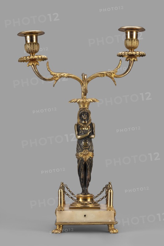 Two-candle candelabra depicting Egyptian women, early 19th century, France, unknown locality; lighting; height 35 cm, width 22.5 cm, depth 11.3 cm, A decorative two-armed candlestick featuring a central sculpted female figure with crossed arms, standing on a rectangular base; the piece combines dark metallic and bright gold tones with a light stone-colored base, a group of ornate gold arms extending outward with leaf-like motifs and two candle holders at the ends, a gold floral element at the figure’s waist, small chained railings connecting four gold posts around the base, and gold paw-shaped feet supporting the stone-colored platform.